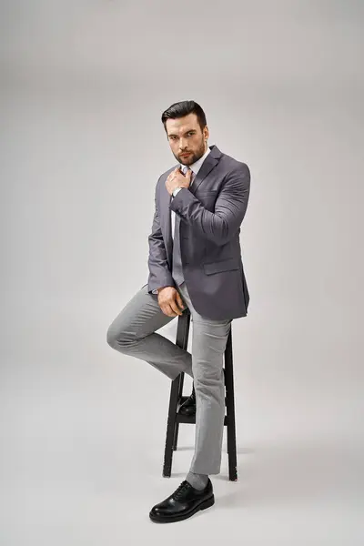 Gutaussehender und selbstbewusster Geschäftsmann in formaler Kleidung, angelehnt an einen hohen Hocker auf grauem Hintergrund, Eleganz — Stockfoto