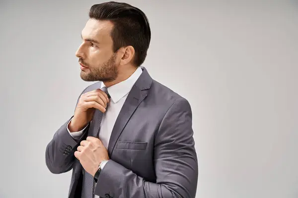 Pensativo hombre de negocios en traje ajustando su corbata sobre fondo gris, concepto de moda corporativa - foto de stock