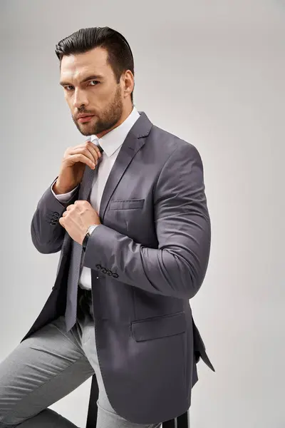 Pensativo hombre de negocios en traje ajustando su corbata sobre fondo gris, concepto de estilo corporativo - foto de stock