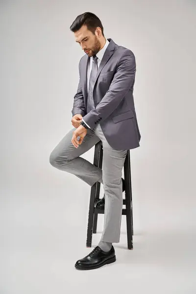 Gutaussehender und selbstbewusster Geschäftsmann im Anzug, gestützt auf einen hohen Hocker auf grauem Hintergrund, Führung — Stockfoto