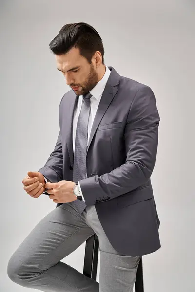 Gutaussehender und selbstbewusster Geschäftsmann im Anzug, der auf einem hohen Hocker auf grauem Hintergrund sitzt, Führung — Stockfoto