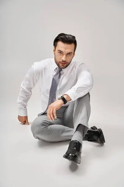 Carismatico e affascinante uomo d'affari in abito formale seduto sul pavimento in studio con sfondo grigio — Foto stock