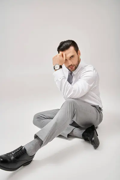 Stylish and charming businessman in formal wear sitting on floor in studio with grey background — Stock Photo