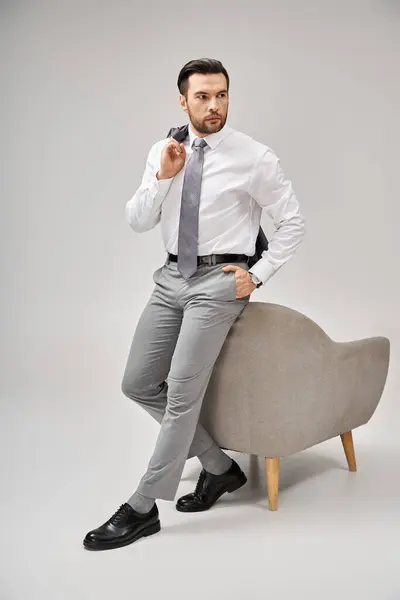 Hombre de negocios guapo en traje posando con la mano en el bolsillo y apoyándose en el sillón sobre fondo gris - foto de stock