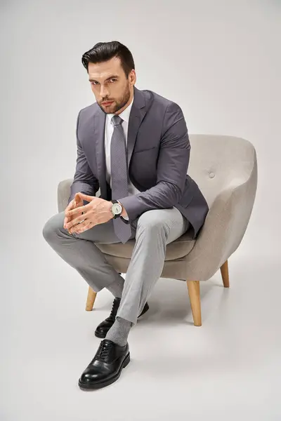 Hombre de negocios seguro y guapo en traje elegante sentado en sillón cómodo sobre fondo gris - foto de stock