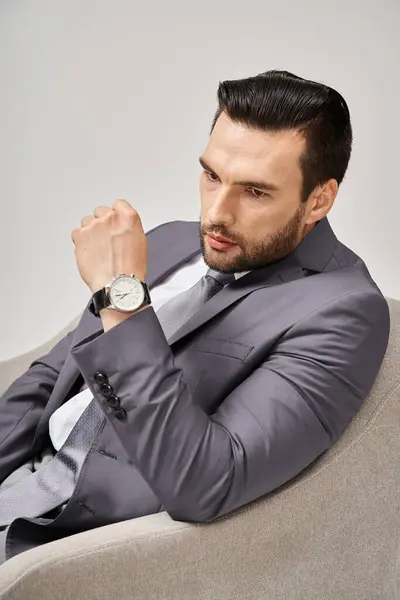 Retrato de empresario pensativo en traje elegante sentado en sillón cómodo sobre fondo gris, 30s - foto de stock