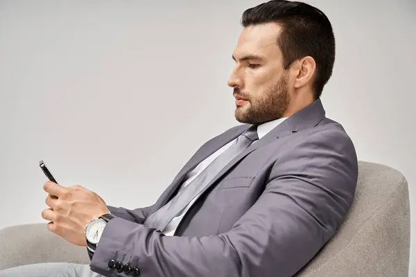 Businessman with bristle sitting on armchair and using smartphone on grey background, social media — Stock Photo