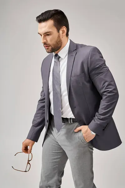 Уравновешенный бизнесмен в формальной одежде держа очки и ходить с рукой в кармане на сером фоне — стоковое фото