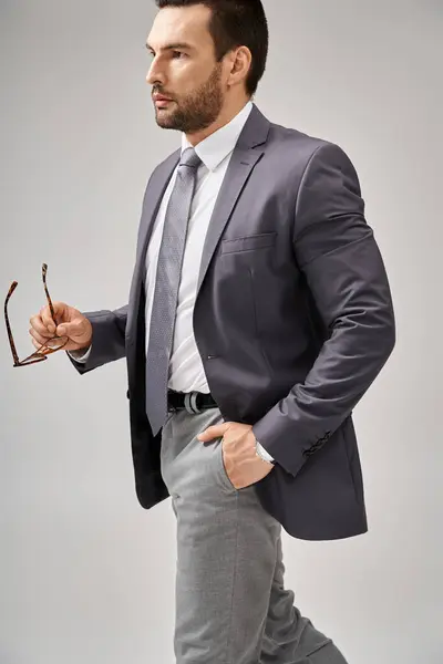 Confident businessman in formal wear holding glasses and standing with hand in pocket on grey — Stock Photo