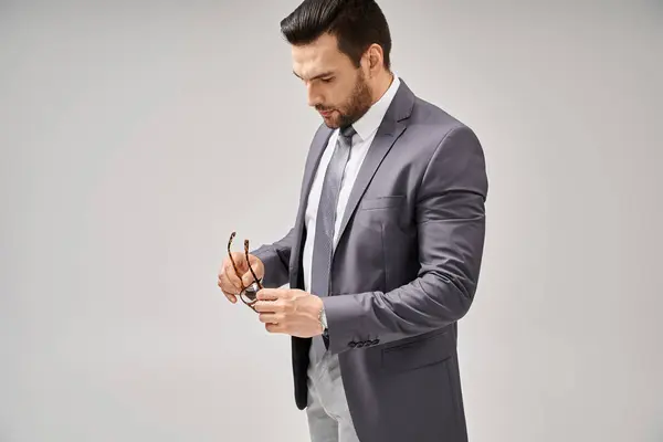 Hombre de negocios guapo en ropa formal sosteniendo gafas y de pie sobre fondo gris, elegancia - foto de stock