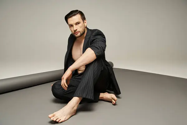 Модний і красивий чоловік з голими грудьми і ногами позує в костюмі з шпильками на сірому фоні — стокове фото