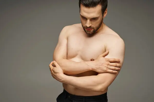 Portrait of brunette and handsome man with bare chest posing and touching muscles on grey backdrop — Stock Photo