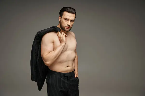 Mann in seinen Dreißigern posiert mit nackter Brust im Nadelstreifenanzug und hält Blazer auf grau, Hand in Tasche — Stockfoto