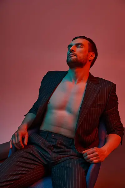 Handsome man with bare chest posing in pinstripe suit and sitting in studio with red and blue light — Stock Photo
