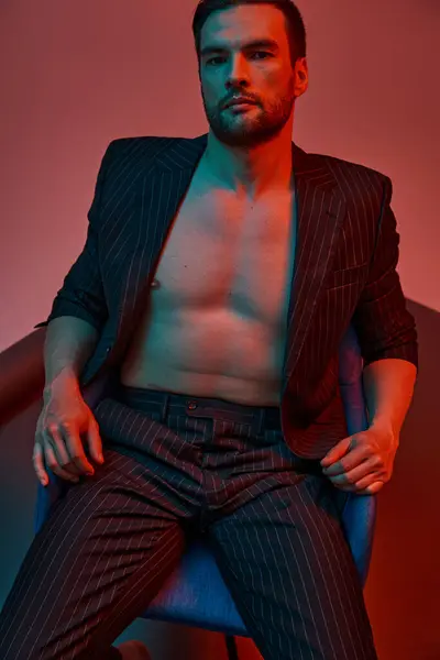 Brunette man with bare chest posing in pinstripe suit and sitting in studio with red and blue light — Stock Photo
