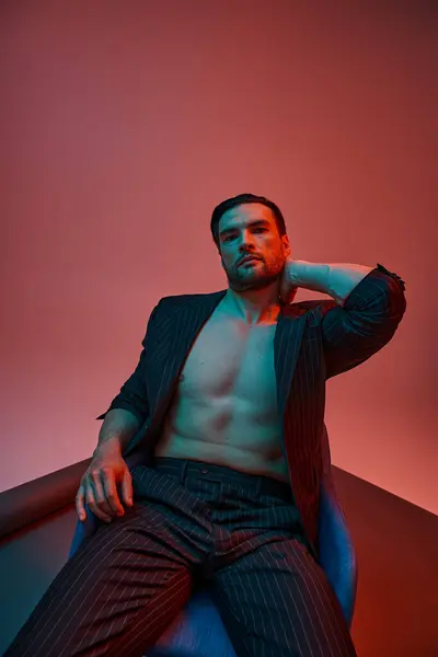 Man with bare chest posing in elegant pinstripe suit and sitting in studio with red and blue light — Stock Photo