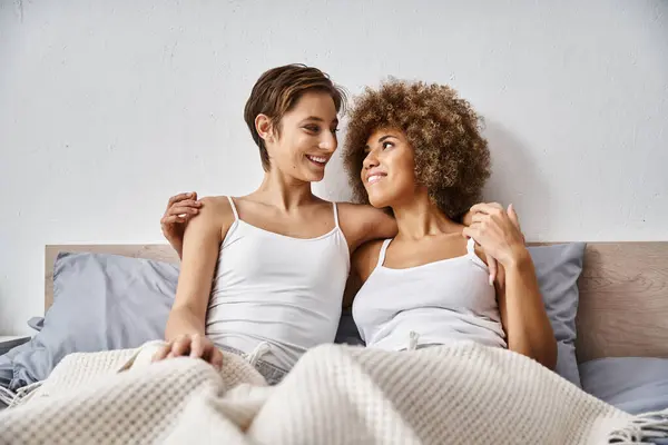 Happy and multicultural lesbian couple hugging while sitting together on bed, comfort and love — Stock Photo