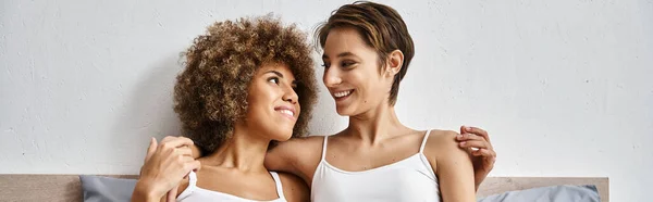Cheerful and multicultural lesbian couple hugging while resting together on bed, horizontal banner — Stock Photo