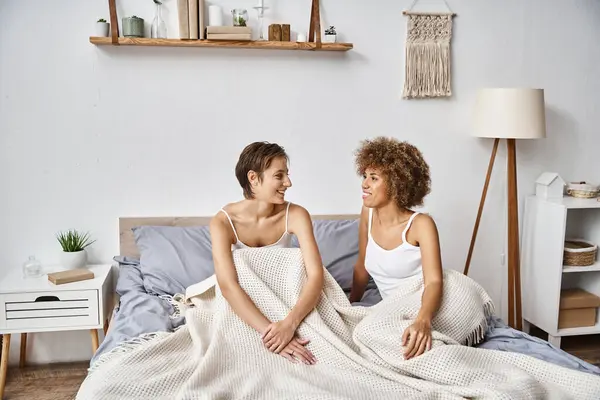 Heureuse femme afro-américaine avec les cheveux bouclés assis avec petite amie sur le lit le matin, confortable — Photo de stock