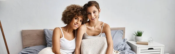 Heureuse femme afro-américaine avec les cheveux bouclés assis avec petite amie sur le lit le matin, bannière — Photo de stock