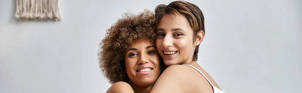 Happy multicultural lesbian couple hugging each other and looking at camera in bedroom, banner — Stock Photo