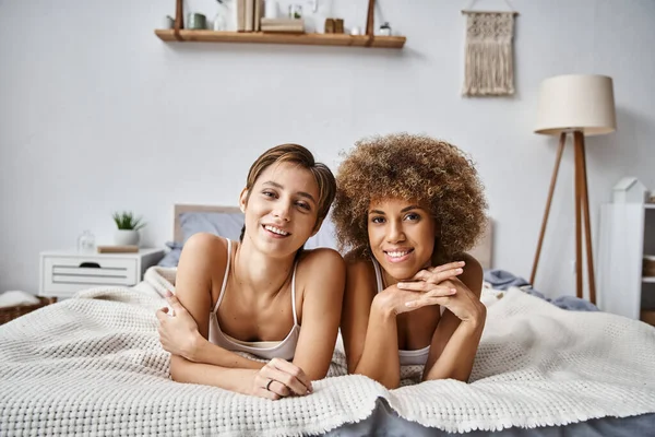 Счастливые лесбийские партнеры смотрят в камеру, лежа вместе на кровати дома, уютные моменты — стоковое фото