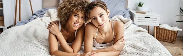 Радісні багатокультурні лесбійські партнери дивляться на камеру, лежачи разом на ліжку вдома, банер — стокове фото