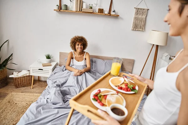 Felice donna afroamericana guardando fidanzata vassoio con frittelle, colazione a letto — Foto stock