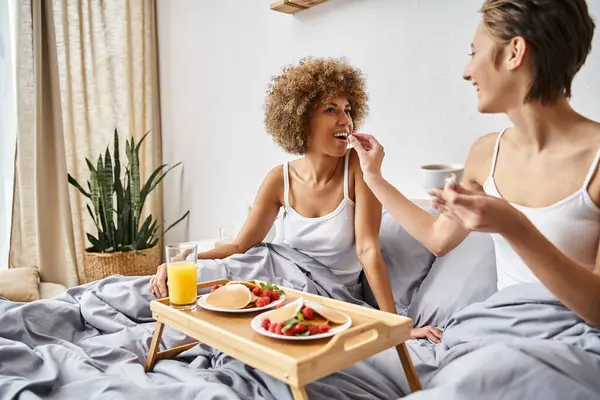 Happy woman feeding her african american girlfriend with raspberry while having breakfast in bed — Stock Photo