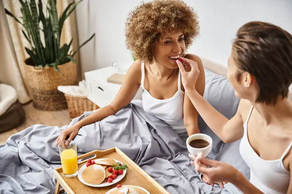 Mulher alegre alimentando sua namorada afro-americana com framboesa enquanto toma café da manhã na cama — Fotografia de Stock