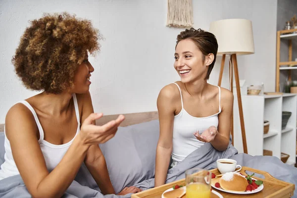 Cheerful multicultural lesbian couple in pajamas gesturing and chatting near breakfast in bed — Stock Photo