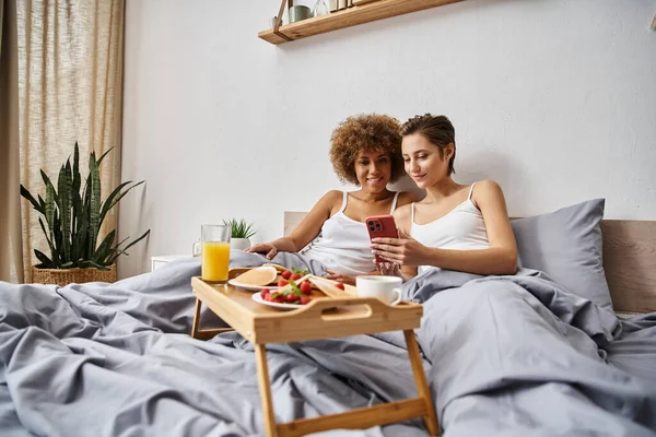Alegre pareja lésbica multicultural en pijama usando smartphone cerca del desayuno en bandeja en la cama - foto de stock