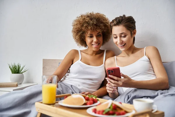 Feliz casal lésbico multicultural em pijama usando smartphone perto do café da manhã na bandeja na cama — Fotografia de Stock