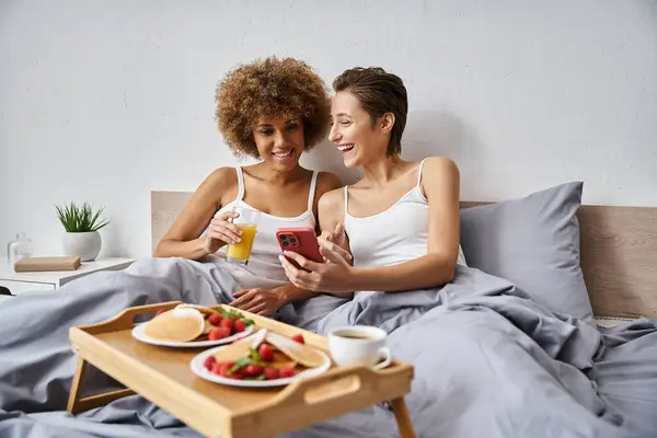 Felice donna lesbica in pigiama ridere e utilizzando smartphone vicino fidanzata afro-americana — Foto stock