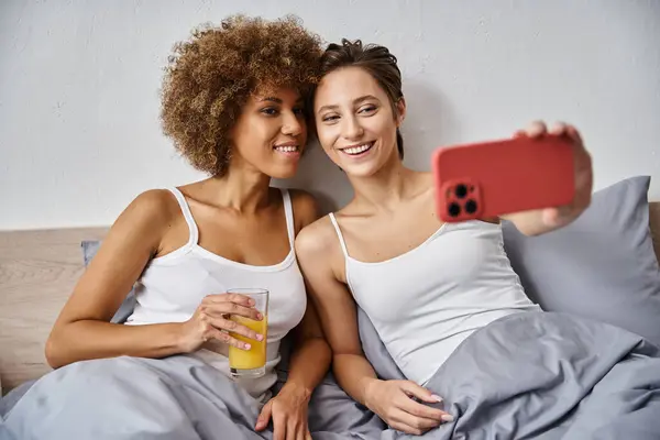 Femme heureuse prenant selfie sur smartphone près de petite amie afro-américaine avec verre de jus d'orange — Photo de stock