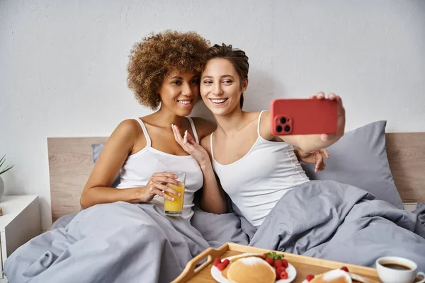 Happy woman taking selfie on smartphone with african american girlfriend near breakfast tray — Stock Photo