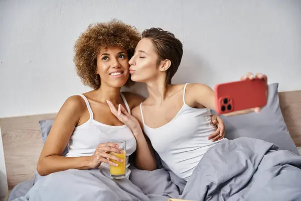 Happy woman taking selfie while kissing african american girlfriend with glass of orange juice — Stock Photo