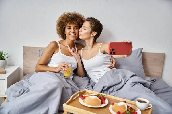 Happy woman taking selfie while kissing african american girlfriend near breakfast tray on bed — Stock Photo