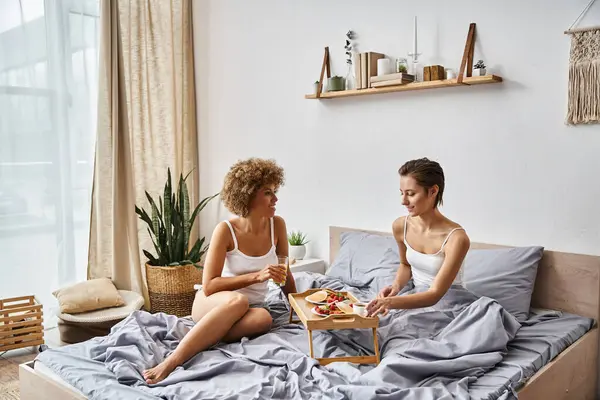 Fröhliches und junges multikulturelles lesbisches Paar im Schlafanzug plaudert neben Tablett mit Frühstück im Bett — Stockfoto