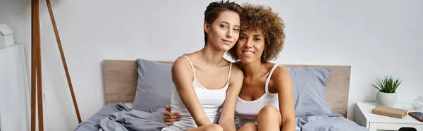 Heureux et jeune couple lesbienne multiculturel en pyjama assis ensemble sur le lit, bannière horizontale — Photo de stock