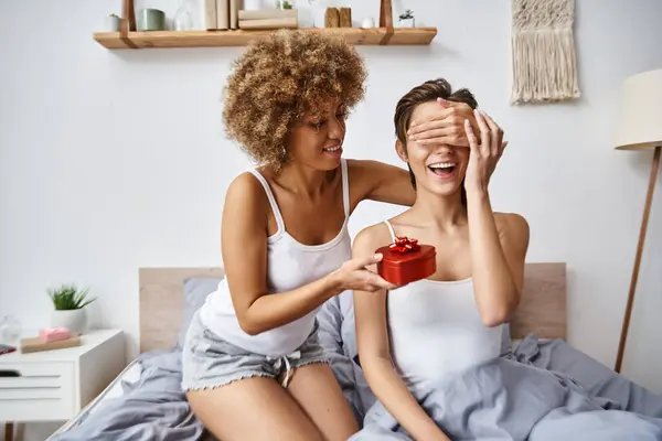 Afro-américaine femme tenant présent rouge et couvrant les yeux de petite amie heureuse le jour de la Saint-Valentin — Photo de stock