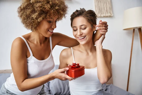 Heureuse femme afro-américaine tenant cadeau rouge près de copine excitée en pyjama le jour de la Saint-Valentin — Photo de stock