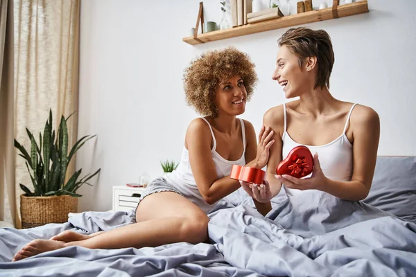 Donna allegra in pigiama apertura scatola regalo a forma di cuore vicino fidanzata afro-americana in camera da letto — Foto stock