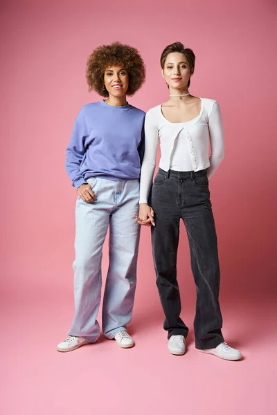 Happy multicultural lesbian women holding hands while standing on pink background, lgbtq couple — Stock Photo