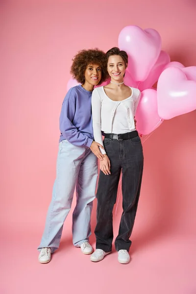 Concepto de San Valentín, alegre pareja lesbiana multicultural sonriendo cerca de globos en forma de corazón - foto de stock