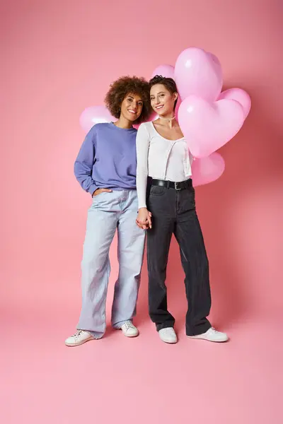 Concepto de San Valentín, pareja lesbiana multicultural positiva sonriendo cerca de globos en forma de corazón - foto de stock
