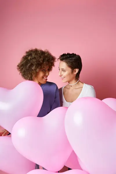Concepto de San Valentín, feliz pareja lesbiana interracial sonriendo cerca de globos en forma de corazón rosa - foto de stock