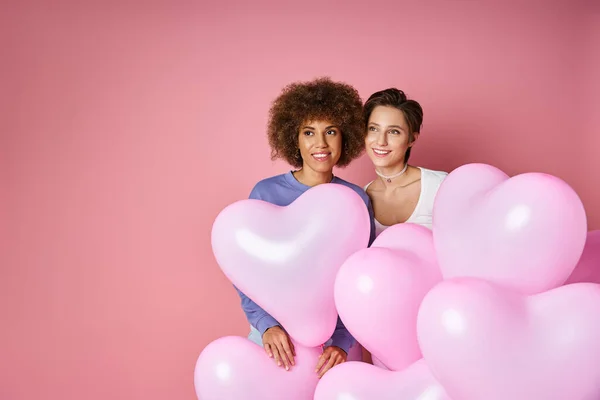 Valentinstag-Konzept: verträumtes lesbisches Paar lächelt neben rosafarbenen herzförmigen Luftballons — Stockfoto
