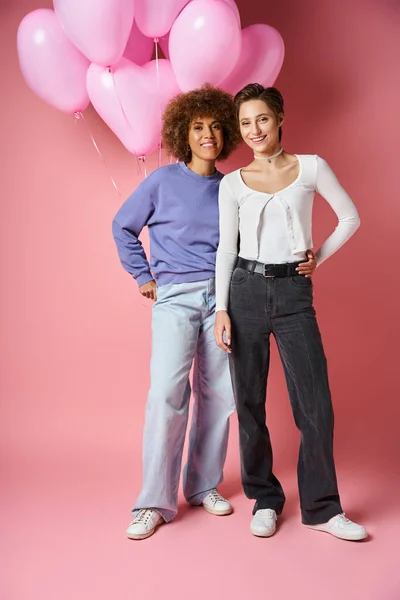 Щаслива міжрасова лесбіянка пара посміхається біля рожевих кульок у формі серця, день Святого Валентина — стокове фото