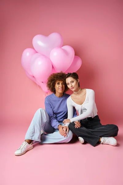 Glückliches multikulturelles lesbisches Paar, das neben rosa herzförmigen Luftballons sitzt, Valentinstag — Stockfoto